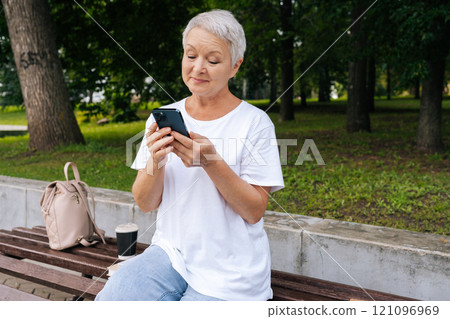 Focused beautiful elderly woman learning to use modern smartphone. Gray-haired grandma sitting on bench in city park, typing on mobile phone. Pensioner surfing Internet with cellphone. Focused beautiful elderly woman learning to use modern smartphone. Gray-haired grandma sitting on bench in city park, typing on mobile phone. Pensioner surfing Internet with cellphone. 121096969