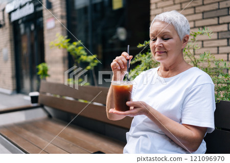 Waist up portrait of serene senior woman enjoying iced coffee sitting on bench outside city cafe, contently stirring with straw on sunny summer day, capturing urban lifestyle for mature adults. Waist up portrait of serene senior woman enjoying iced coffee sitting on bench outside city cafe, contently stirring with straw on sunny summer day, capturing urban lifestyle for mature adults. 121096970