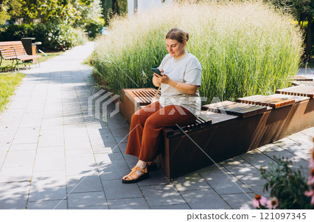 Happy woman chatting on phone and relaxing on the bench outdoors. Urban people concept. Smiling woman text messaging on smart phone at sunny day, full body 121097345