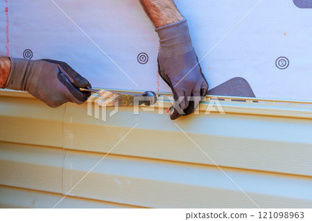 Worker carefully installs vinyl siding on house, ensuring level alignment while using hammer tools. Worker carefully installs vinyl siding on house, ensuring level alignment while using hammer tools. 121098963