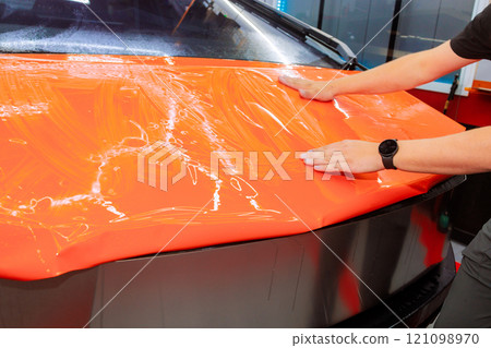Person is smoothing out an orange vinyl wrap on car hood in workshop, ensuring bubble free finish for protect body automotive 121098970