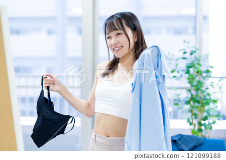 Smiling young woman choosing clothes and underwear in front of a full-length mirror, choosing sports bra and underwear 121099188