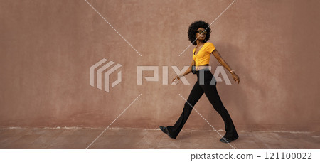 Stylish individual confidently walks against a textured brown wall in a vibrant yellow top and black pants 121100022