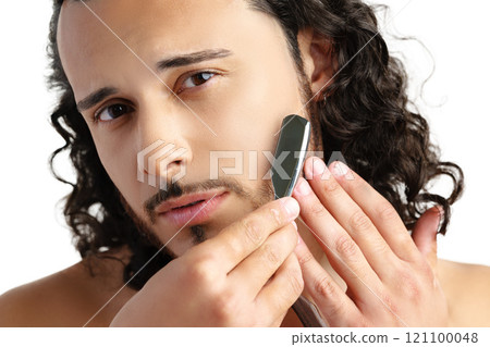 Man with curly hair grooming his beard carefully using a straight razor against a plain white background 121100048