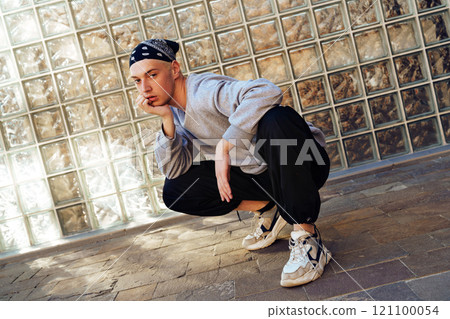 A young man wearing a gray sweatshirt and black pants squats in front of a glass block wall A young man wearing a gray sweatshirt and black pants squats in front of a glass block wall 121100054