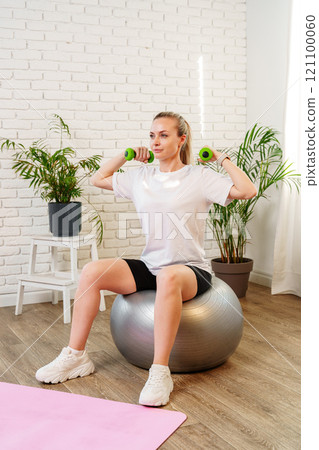 Woman exercising with dumbbells on a stability ball in a indoor space 121100060