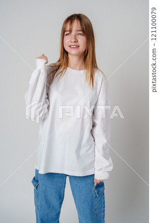 A joyful young girl with long hair wearing a white sweater stands against a plain background A joyful young girl with long hair wearing a white sweater stands against a plain background 121100079