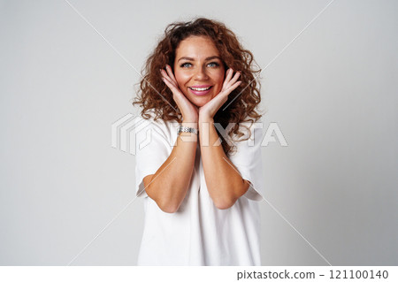 Woman with curly hair smiles joyfully while posing with hands on her cheeks against a light background Woman with curly hair smiles joyfully while posing with hands on her cheeks against a light background 121100140