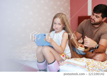 A young girl plays on a tablet while her father brushes her long hair A young girl plays on a tablet while her father brushes her long hair 121100142