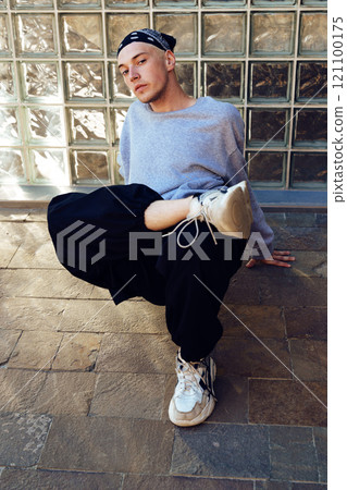 A young man wearing a bandana sits on a tiled floor in front of a glass block wall, striking a pose while wearing grey sweatshirt and black pants A young man wearing a bandana sits on a tiled floor in front of a glass block wall, striking a pose while wearing grey sweatshirt and black pants 121100175