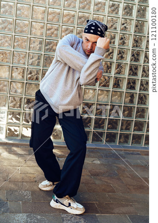 A young man in a grey sweatshirt, black pants, and a bandana poses in front of a glass block wall 121100180