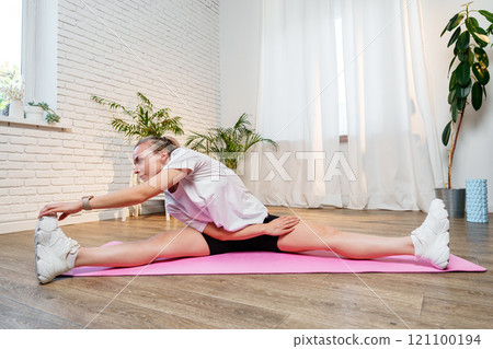 A young woman stretches on a pink mat in a bright room 121100194