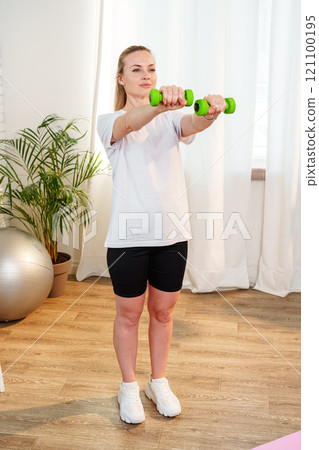 A woman exercises with green dumbbells at home in a bright room 121100195