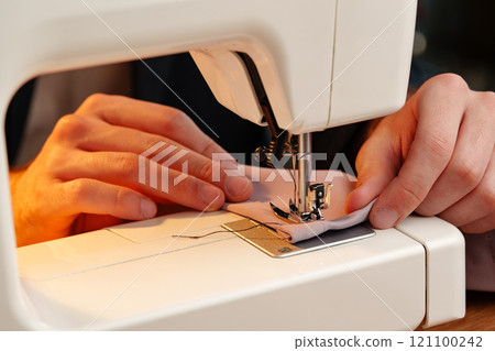 Hands sewing fabric using a sewing machine on a wooden table in a well-lit workspace 121100242