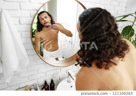 A man with curly hair grooming himself in the bathroom while looking into a round mirror A man with curly hair grooming himself in the bathroom while looking into a round mirror 121100263