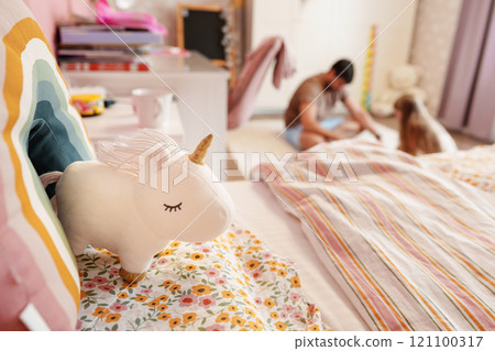 A white unicorn plush toy sits on a bed with a rainbow pillow in a bedroom, blurred figures in the background A white unicorn plush toy sits on a bed with a rainbow pillow in a bedroom, blurred figures in the background 121100317