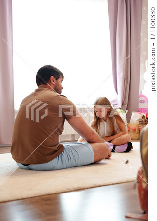 A father and his daughter sit on a rug in their home, the father looking at his daughter with a gentle expression A father and his daughter sit on a rug in their home, the father looking at his daughter with a gentle expression 121100330