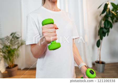 A woman exercises with green dumbbells at home in a bright room A woman exercises with green dumbbells at home in a bright room 121100338