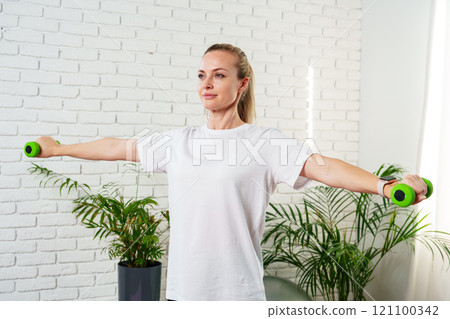 A woman exercises with green dumbbells at home in a bright room A woman exercises with green dumbbells at home in a bright room 121100342