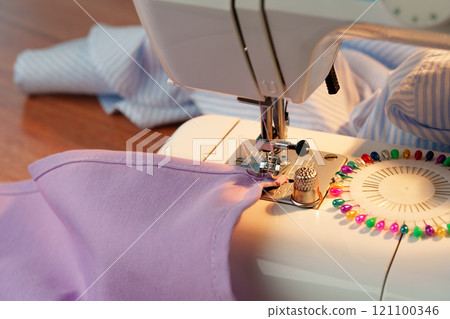 Sewing machine in use on a wooden table, stitching a light fabric in a well-lit indoor workspace Sewing machine in use on a wooden table, stitching a light fabric in a well-lit indoor workspace 121100346