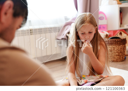 A young girl sits on the floor in a living room, holding a small object in her hand and looking down at it A young girl sits on the floor in a living room, holding a small object in her hand and looking down at it 121100363