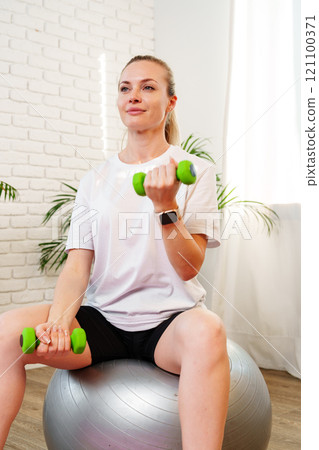 Woman exercising with dumbbells on a stability ball in a indoor space Woman exercising with dumbbells on a stability ball in a indoor space 121100371