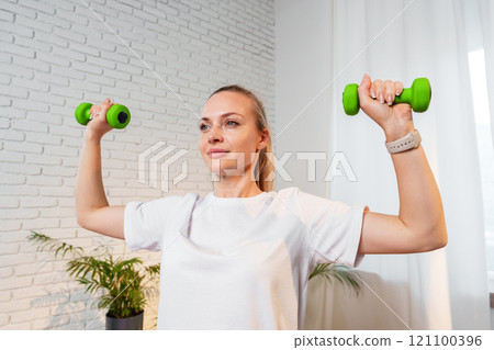 A woman exercises with green dumbbells at home in a bright room A woman exercises with green dumbbells at home in a bright room 121100396