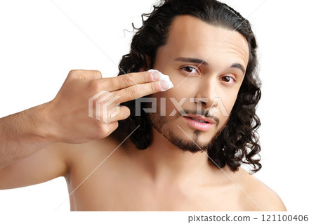 A young man applying skincare cream on his face with a focused expression, demonstrating personal grooming at home 121100406