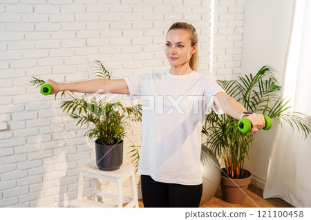 A woman exercises with green dumbbells at home in a bright room A woman exercises with green dumbbells at home in a bright room 121100458
