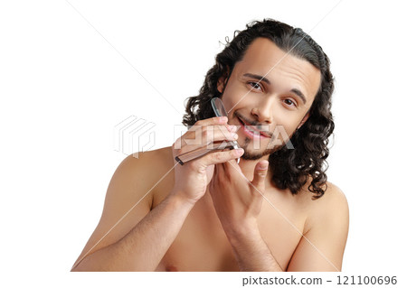 A man with curly hair grooming his beard in a well-lit space while using a straight razor 121100696