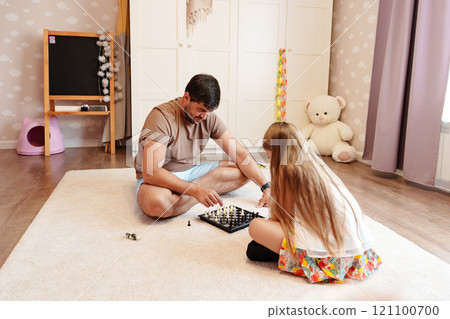 A father and daughter play chess on a white rug in a home 121100700
