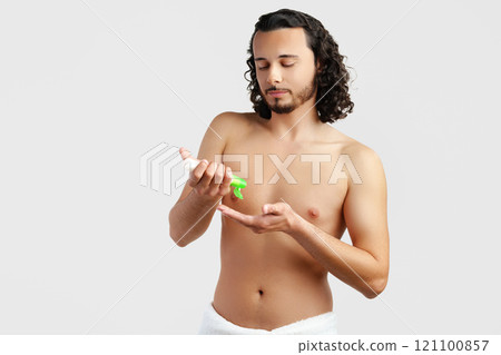 A young man applying skincare cream on his face with a focused expression, demonstrating personal grooming at home 121100857