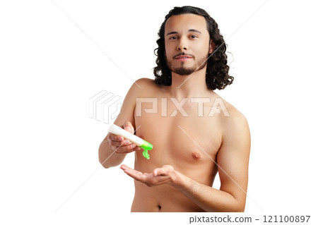 A young man applying skincare cream on his face with a focused expression, demonstrating personal grooming at home 121100897