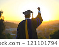 A proud graduate raises a fist in celebration against the backdrop of a vibrant sunset. 121101794