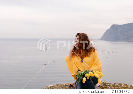 Portrait happy woman woman with long hair against a background of mountains and sea. Holding a bouquet of yellow tulips in her hands, wearing a yellow sweater Portrait happy woman woman with long hair against a background of mountains and sea. Holding a bouquet of yellow tulips in her hands, wearing a yellow sweater 121104772