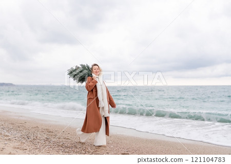 Blond woman Christmas tree sea. Christmas portrait of a happy woman walking along the beach and holding a Christmas tree on her shoulder. She is wearing a brown coat and a white suit. Blond woman Christmas tree sea. Christmas portrait of a happy woman walking along the beach and holding a Christmas tree on her shoulder. She is wearing a brown coat and a white suit. 121104783