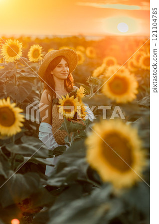 A girl in a hat on a beautiful field of sunflowers against the sky in the evening light of a summer sunset. Sunbeams through the flower field. Natural background. 121104785