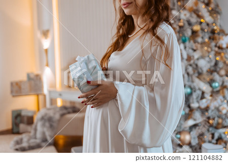 Pregnant woman holds a gift on her stomach against Christmas tree with lights. family holiday concept Christmas holidays. 121104882
