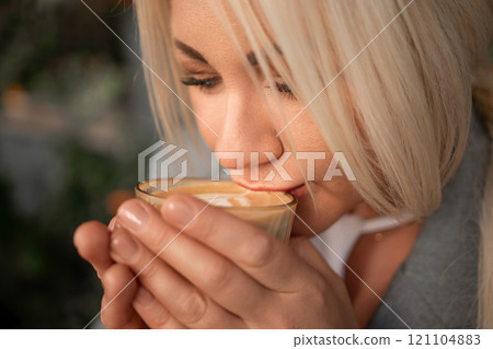 A blonde woman is drinking coffee from a glass. She is smiling and she is enjoying her drink. 121104883