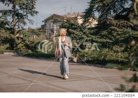 A woman is walking down a sidewalk with a cup in her hand. She is wearing a jacket and jeans. A woman is walking down a sidewalk with a cup in her hand. She is wearing a jacket and jeans. 121104884