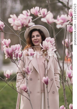 Woman magnolia flowers, surrounded by blossoming trees., hair down, white hat, wearing a light coat. Captured during spring, showcasing natural beauty and seasonal change. Woman magnolia flowers, surrounded by blossoming trees., hair down, white hat, wearing a light coat. Captured during spring, showcasing natural beauty and seasonal change. 121105523