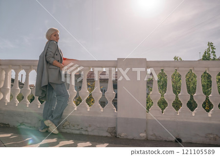 A woman in a gray jacket and blue jeans is leaning against a white railing. She is holding a book in her hand. The scene has a calm and relaxed mood, with the woman enjoying a quiet moment outdoors. 121105589