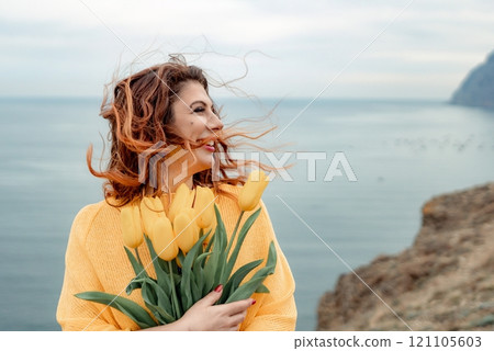 Portrait of a happy woman with hair flying in the wind against the backdrop of mountains and sea. Holding a bouquet of yellow tulips in her hands, wearing a yellow sweater 121105603