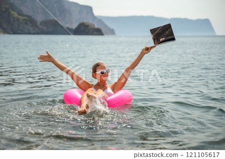 A woman is floating in a pink inflatable raft with a laptop in her hand. She is smiling and she is enjoying her time in the water. 121105617