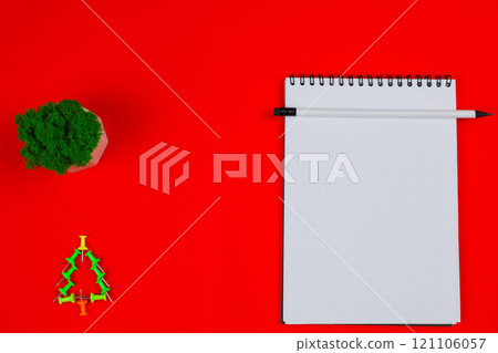 A white notebook with a black spring lies in the center of the photo, a white pencil with a black lead and an eraser lies on top, on the left there is a green plant and a green pushpin Christmas tree A white notebook with a black spring lies in the center of the photo, a white pencil with a black lead and an eraser lies on top, on the left there is a green plant and a green pushpin Christmas tree 121106057