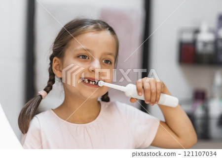 Little Caucasian girl with missing baby teeth brushing teeth with electric toothbrush in bathroom. Child looking into mirror while practicing oral hygiene and dental care. Little Caucasian girl with missing baby teeth brushing teeth with electric toothbrush in bathroom. Child looking into mirror while practicing oral hygiene and dental care. 121107034