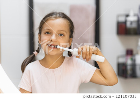 Little Caucasian girl with missing front teeth brushing teeth with electric toothbrush, smiling in bathroom mirror, building healthy habits. 121107035