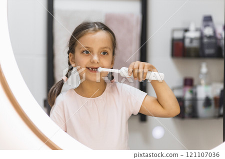 Little Caucasian girl with lost baby teeth using electric toothbrush, looking in bathroom mirror, promoting dental hygiene. Little Caucasian girl with lost baby teeth using electric toothbrush, looking in bathroom mirror, promoting dental hygiene. 121107036
