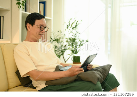 Senior man sitting on couch using a tablet to read or browse in a bright living room 121107299