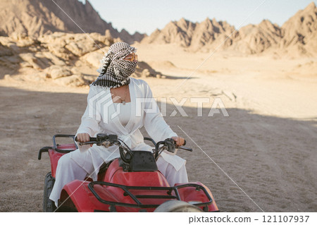 A rather attractive woman is going on a safari, driving an ATV in the desert. Beautiful young woman with a scarf on her head sits on a red four-wheeled ATV and rides towards adventure 121107937